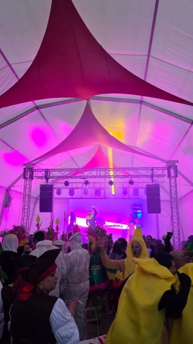 Fiesta en carpa con luces moradas y público bailando en atuendos coloridos.