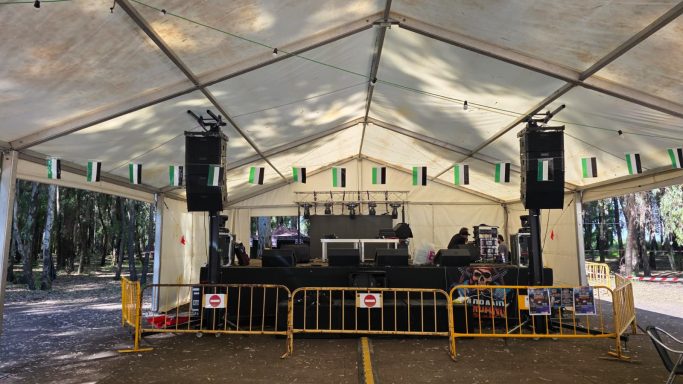 Escenario montado bajo una carpa con altavoces y barreras de seguridad, rodeado de árboles.