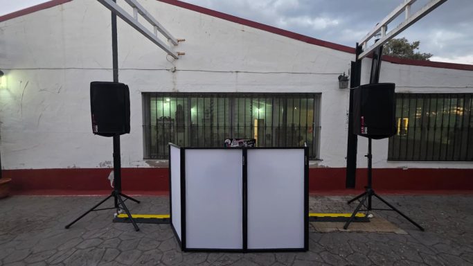 Escenario con una mesa central y altavoces a los lados, frente a un edificio.