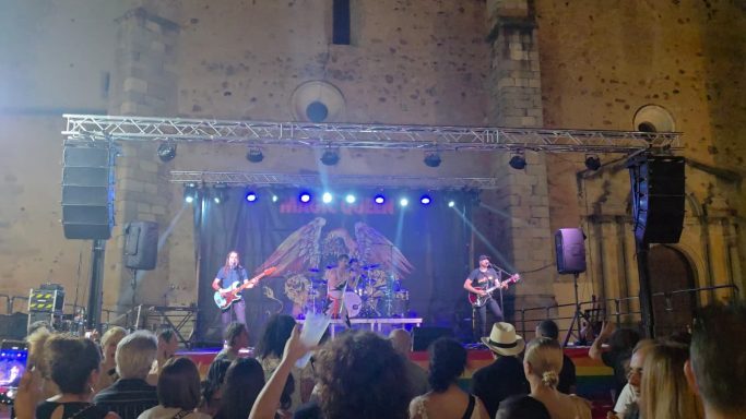 Concierto en un escenario frente a un público, con luces coloridas y una iglesia antigua de fondo.