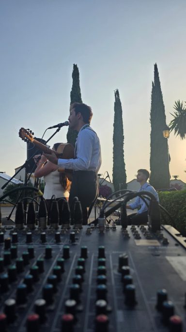 Músicos tocando en un evento al aire libre, con árboles altos de fondo.