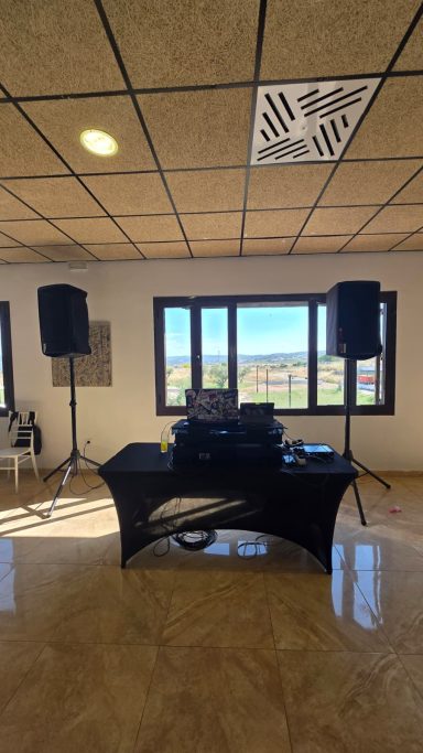 Mesa de DJ con altavoces en una sala con ventanas que muestran un paisaje exterior.